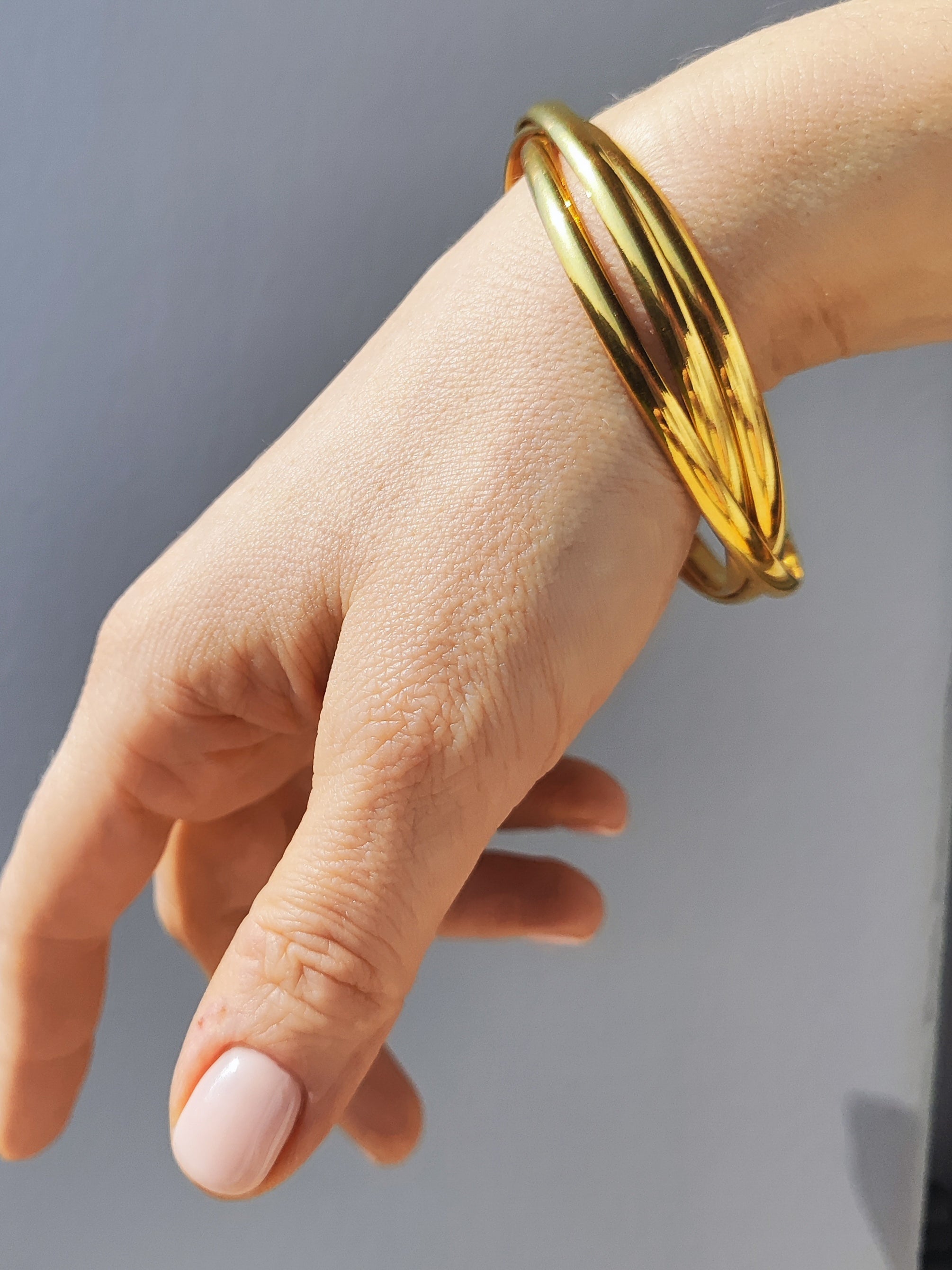 Bracciale Paola in acciaio inossidabile color oro. È ripreso indossato e si nota l'intreccio dei tre bracciali. La mano che lo porta è di fronte ad uno sfondo grigio.
