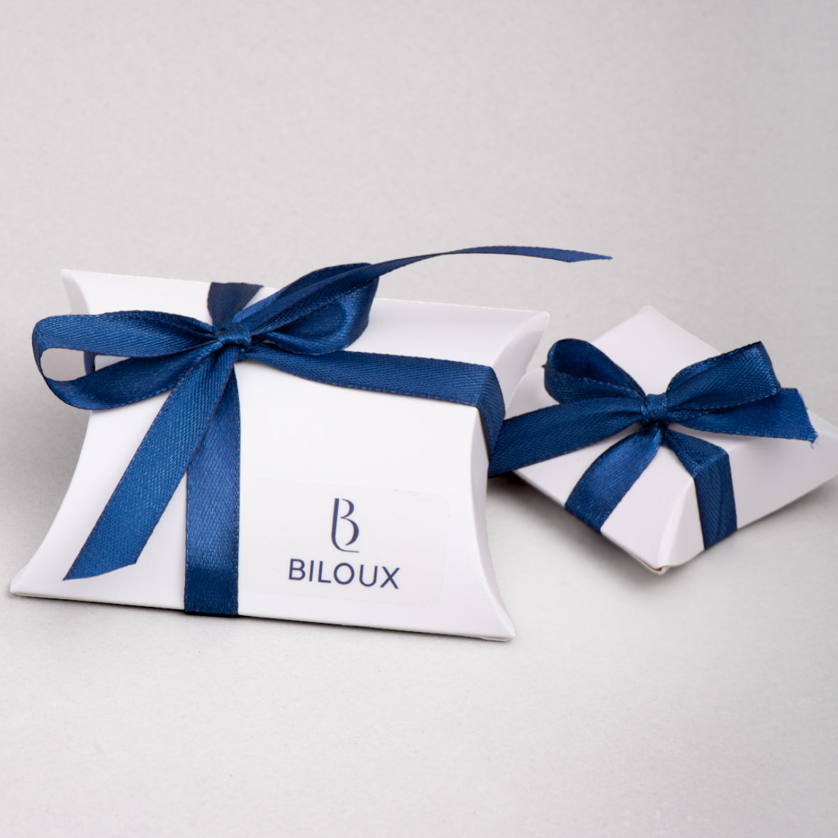 Coppia di pacchetti regalo Biloux in cartoncino bianco con fiocco blu scuro. Sono di due misure diverse, più grande quella a sinistra con logo in evidenza e più piccona quella di destra.