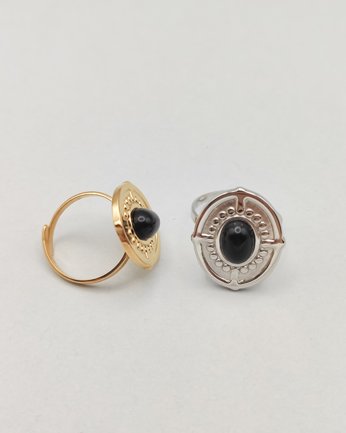 Coppia di anelli Antonella in acciaio inossidabile anallergico color argento e oro con pietra onice. Sono di forma ovale; l'anello di destra, in tinta silver, è rappresentato frontalmente, quello di destra in gold è poggiato su un lato per evidenziare la chiusura regolabile. Si vede la pietra onice al centro, contornata da una lavorazione a pallini in rilievo, così come il bordo dell'anello e dei trattini che riprendono i quatto punti cardinali.