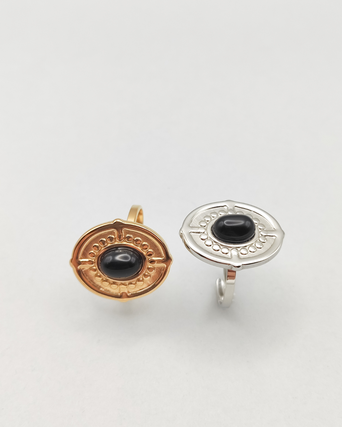 Coppia di anelli Antonella in acciaio inossidabile anallergico color oro e argento con pietra onice. Sono di forma ovale, l'anello di sinistra in tinta gold è ripreso frontalmente mentre quello di destra, in silver, e messo lateralmente verso l'alto. Si vede la pietra onice al centro, contornata da una lavorazione a pallini in rilievo, così come il bordo dell'anello e dei trattini che riprendono i quatto punti cardinali.