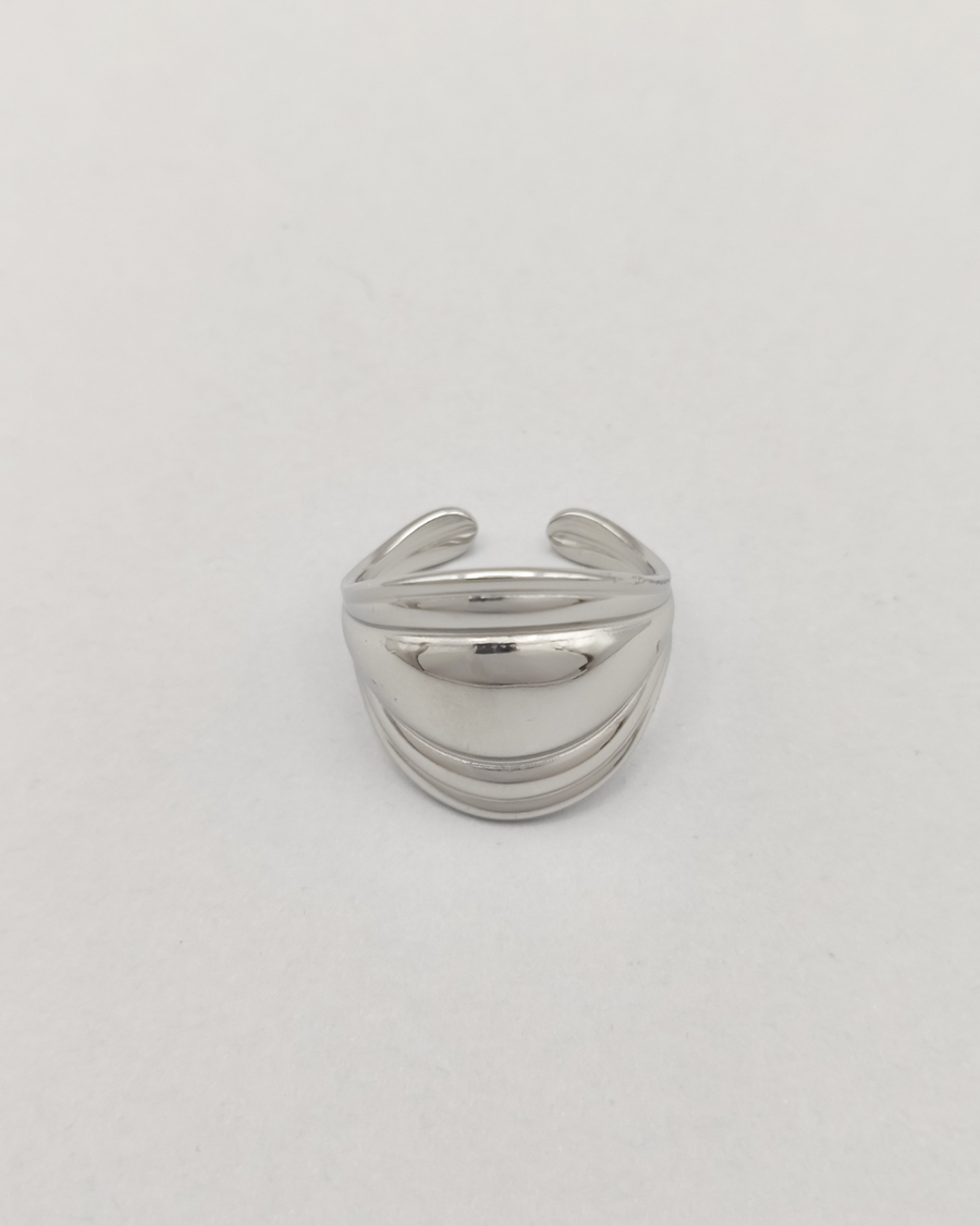 Anello Carlotta in acciaio inossidabile anallergico color argento. È rappresentato frontalmente su uno sfondo grigio chiaro. Ha una forma bombata al centro che si ristringe verso i lati ed ha un finish lucido e si vede la chiusura regolabile sul retro