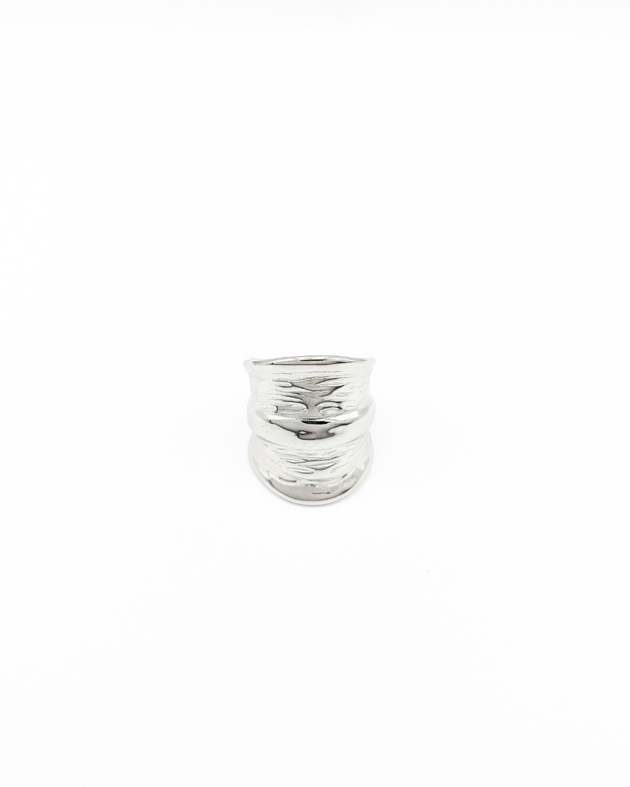 Anello Odine in acciaio inossidabile color argento. È fotografato frontalmente e lascia vedere bene la forma più larga al centro che si restringe verso il retro. Ha una fascia più sporgente al centro mentre verso le estremità il design è drappeggiato.