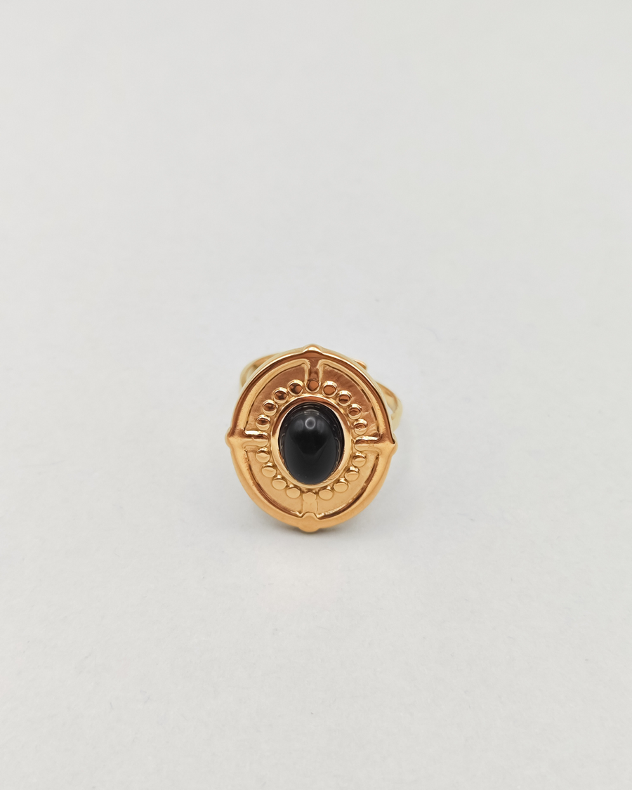 Anello Antonella in acciaio inossidabile anallergico color oro e pietra onice. È di forma ovale e rappresentato frontalmente. Si vede la pietra onice al centro, contornata da una lavorazione a pallini in rilievo, così come il bordo dell'anello e dei trattini che riprendono i quatto punti cardinali.