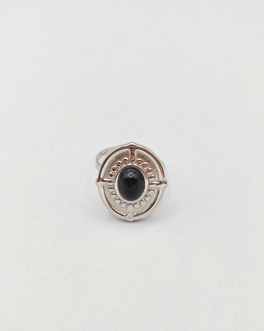 Anello Antonella in acciaio inossidabile anallergico color argento e pietra onice. È di forma ovale e rappresentato frontalmente. Si vede la pietra onice al centro, contornata da una lavorazione a pallini in rilievo, così come il bordo dell'anello e dei trattini che riprendono i quatto punti cardinali.