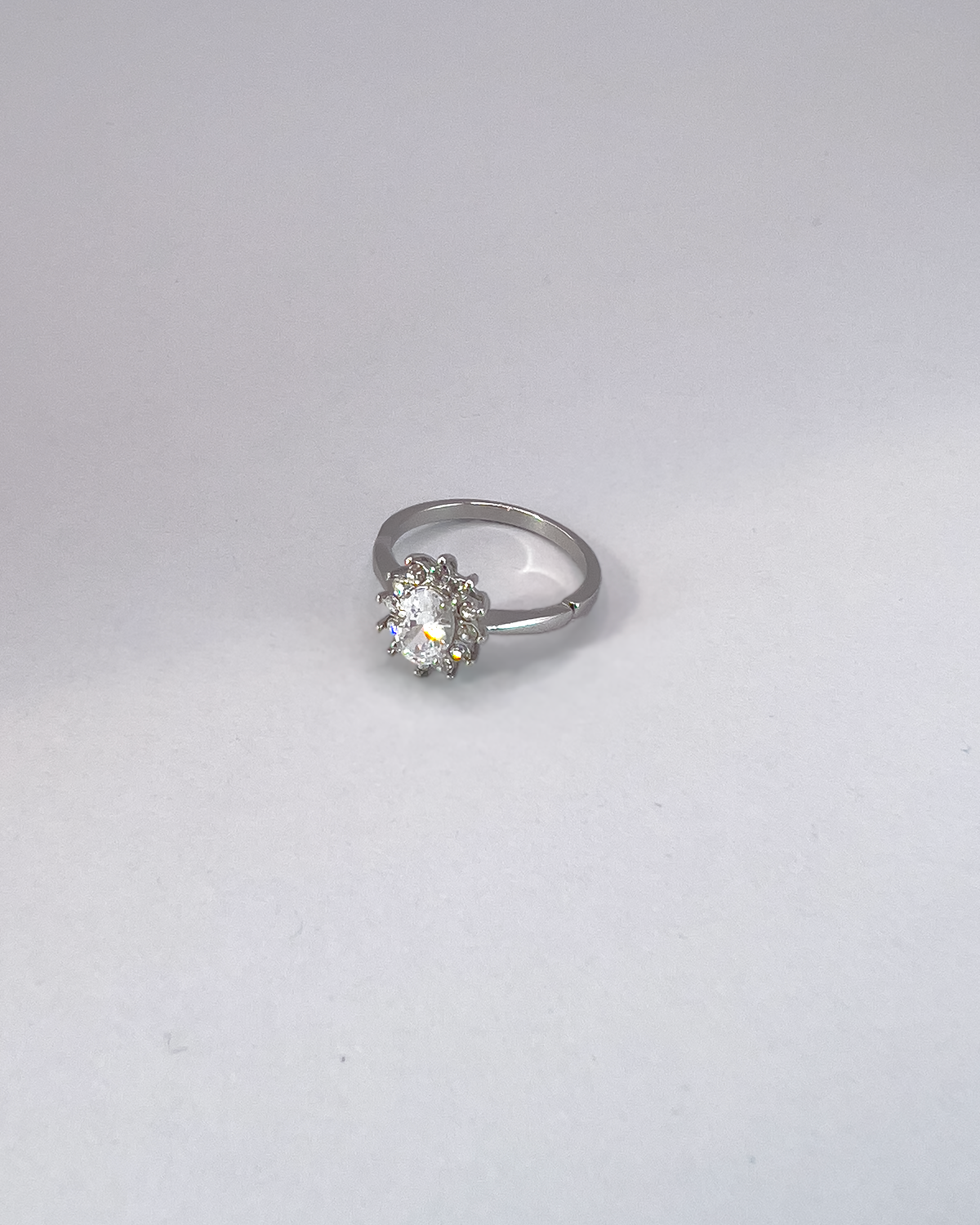 Anello Elisabetta, modello princess, in acciaio inossidabile color argento e zirconi. La pietra centrale è contornata da più pietre piccole. L'anello è ripreso in tre quarti su uno sfondo grigio chiaro.