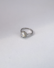 Anello Elisabetta, modello princess, in acciaio inossidabile color argento e zirconi. La pietra centrale è contornata da più pietre piccole. L'anello è ripreso in tre quarti su uno sfondo grigio chiaro.