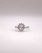 Anello Elisabetta, modello princess, in acciaio inossidabile color argento e zirconi. La pietra centrale è contornata da più pietre piccole. L'anello è ripreso frontalmente su uno sfondo grigio chiaro.