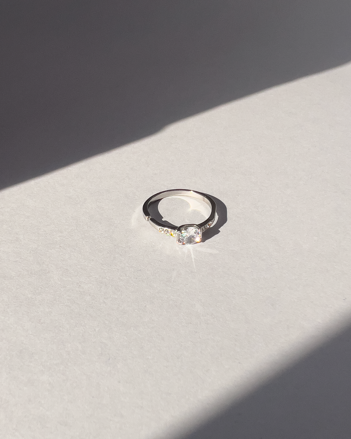 Anello Letizia in acciaio inossidabile anallergico in colore argento e zirconi. La pietra centrale è accompagnata da piccole pietre sui lati. E' rappresentato frontalmente su uno sfondo grigio, ed è illuminato dal sole.