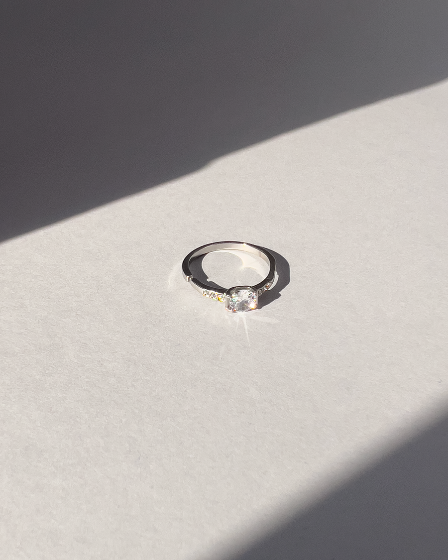 Anello Letizia in acciaio inossidabile anallergico in colore argento e zirconi. La pietra centrale è accompagnata da piccole pietre sui lati. E' rappresentato frontalmente su uno sfondo grigio, ed è illuminato dal sole.