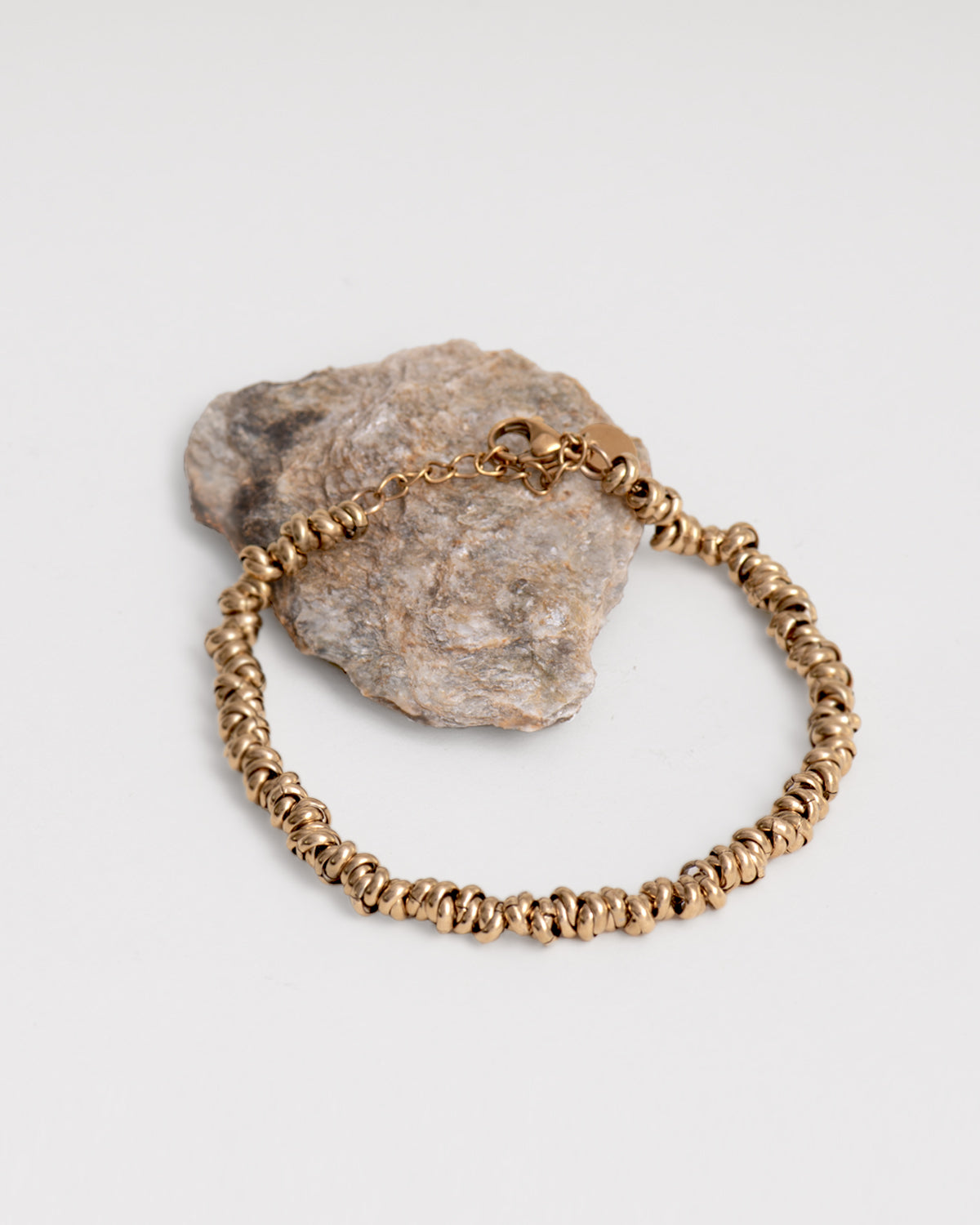 Bracciale Anna in acciaio inossidabile , anallergico e waterproof, color oro. È appoggiato chiuso su una roccia grigia. È composto da tanti nodini infilati in una catenella. La chiusura è a moschettone ed è regolabile in lunghezza.