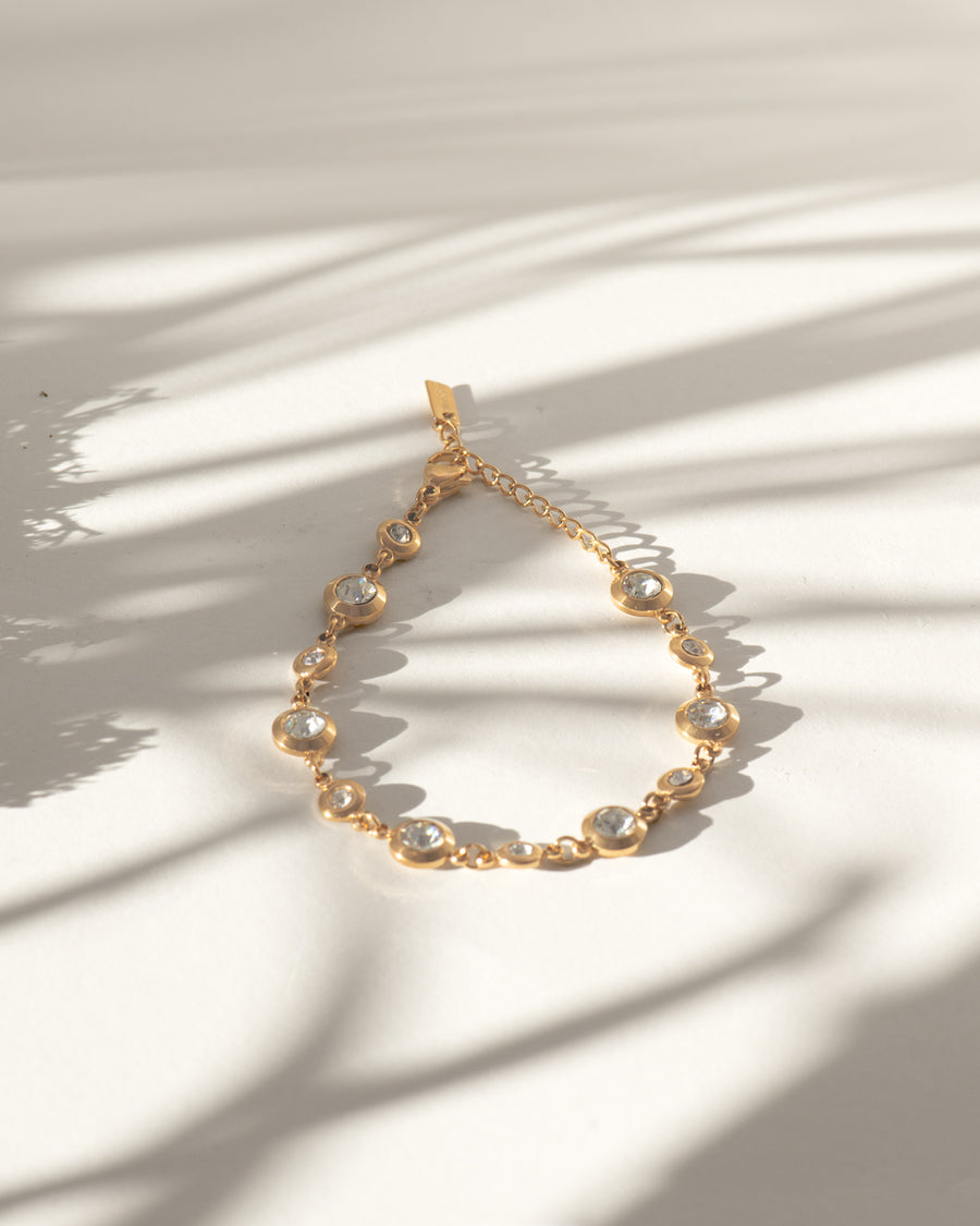Bracciale Chiara in acciaio inossidabile e strass montati su castoni rotondi colore oro. È rappresentato chiuso ed adagiato a goccia su sfondo bianco sul quale si stagliano delle ombre allungate. I castoni alternano una misura grande ad una più piccola e sono distanziati da una catenella. La chiusura è a moschettone e si può regolare la lunghezza. Il materiale è waterproof e nichel free.