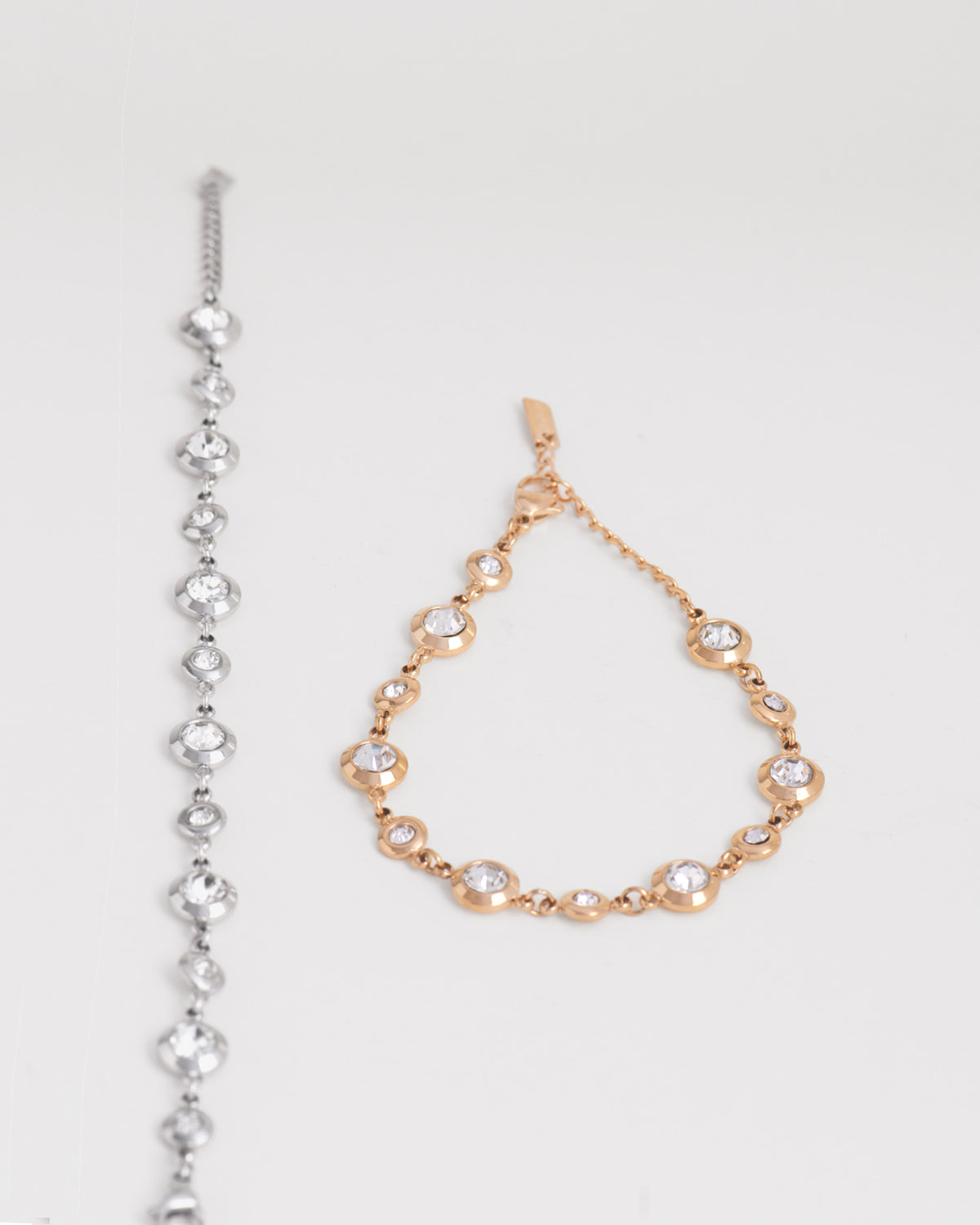 Coppia di bracciali Chiara in acciaio inossidabile e strass montati su castoni rotondi colore argento e oro su sfondo bianco. Il bracciale aragento è raffigurato aperto e disteso, mentre quello oro è chiuso e posizionato a goccia. I castoni alternano una misura grande ad una più piccola e sono distanziati da una catenella. La chiusura è a moschettone e si può regolare la lunghezza. Il materiale è waterproof e nichel free.