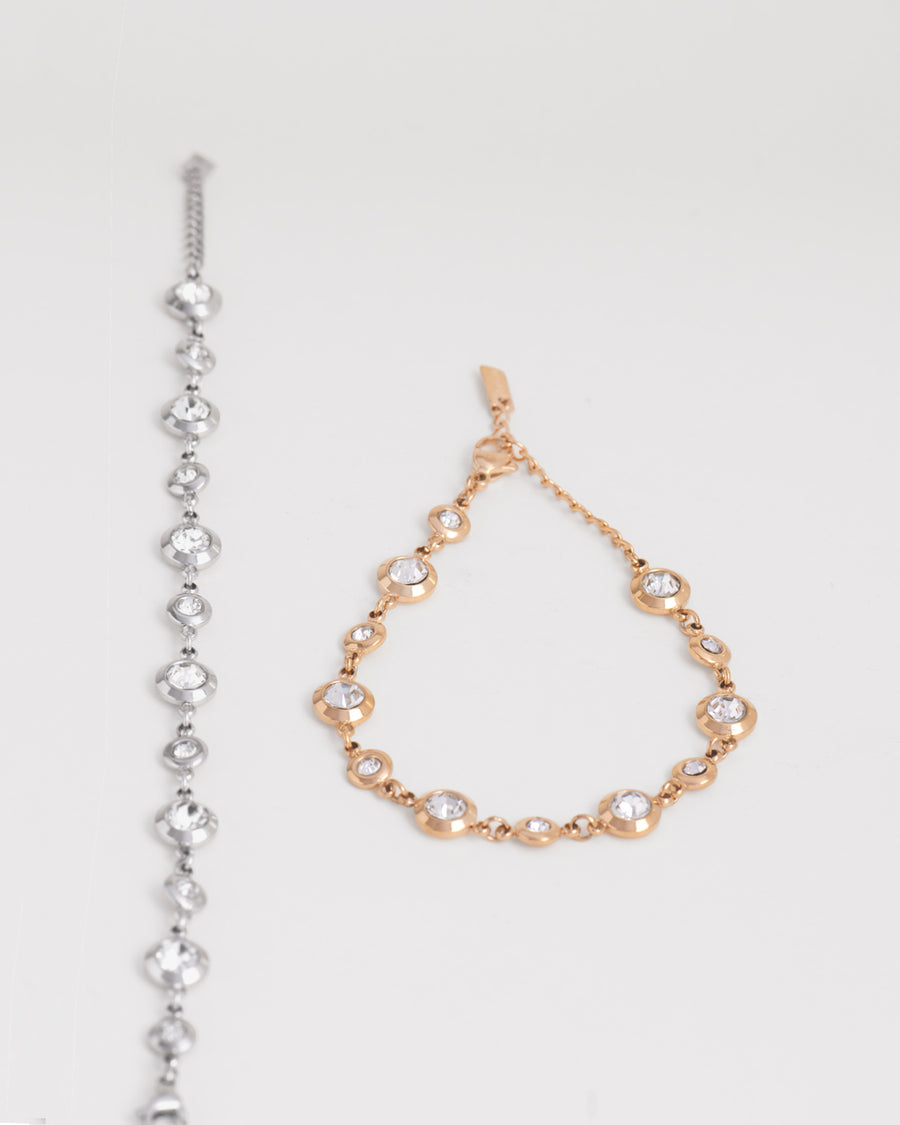 Coppia di bracciali Chiara in acciaio inossidabile e strass montati su castoni rotondi colore argento e oro su sfondo bianco. Il bracciale aragento è raffigurato aperto e disteso, mentre quello oro è chiuso e posizionato a goccia. I castoni alternano una misura grande ad una più piccola e sono distanziati da una catenella. La chiusura è a moschettone e si può regolare la lunghezza. Il materiale è waterproof e nichel free.