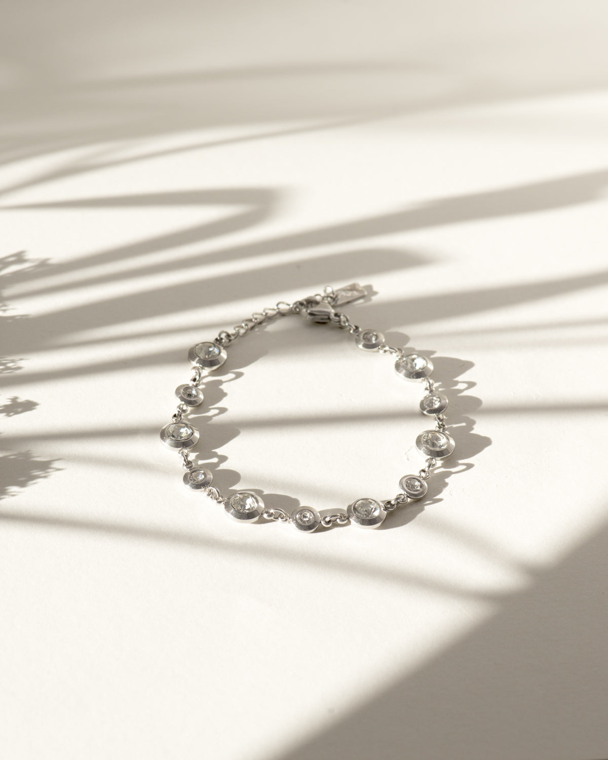 Bracciale Chiara in acciaio inossidabile e strass montati su castoni rotondi colore argento. È rappresentato chiuso ed adagiato su sfondo bianco sul quale si stagliano delle ombre lunghe e sottili. I castoni alternano una misura grande ad una più piccola e sono distanziati da una catenella. La chiusura è a moschettone e si può regolare la lunghezza. Il materiale è waterproof e nichel free.