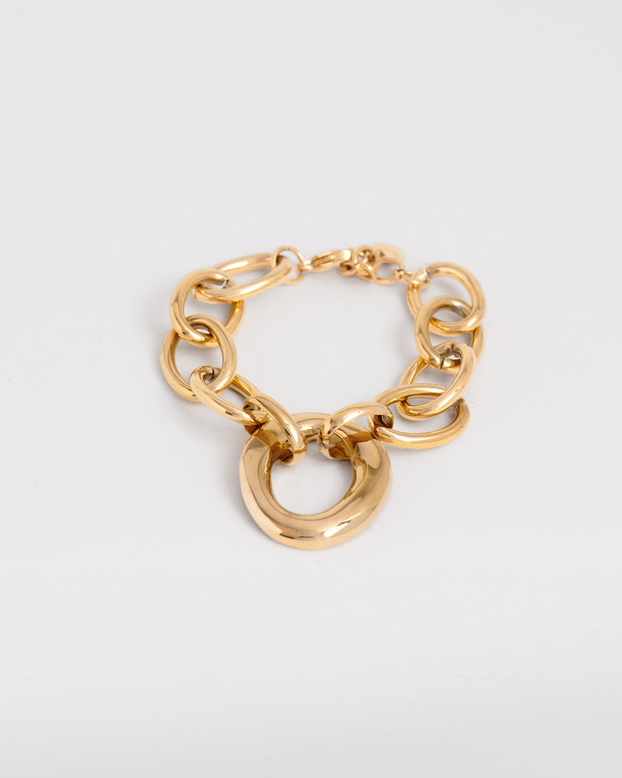 Bracciale Denise in acciaio inossidabile color oro, appoggiato chiuso su uno sfondo bianco. Il bracciale ha una catena a maglie larghe ovali e un ciondolo centrale ovale a sua volta, dalla forma irregolare e finish lucido. La chiusura è a moschettone ed è regolabile. È un bracciale anallergico e resistente all'acqua.