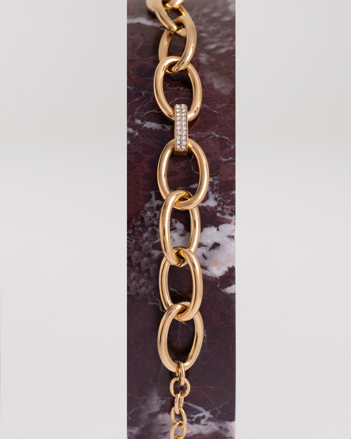 Bracciale Sandra in acciaio inossidabile dorato a maglie larghe ovali con raccordo centrale a doppia fila di strass. È adagiato su un profilo di marmo rosso e si vede la fine della catenella che permette di regolare la lunghezza. Come sempre è in materiale anallergico e waterproof.