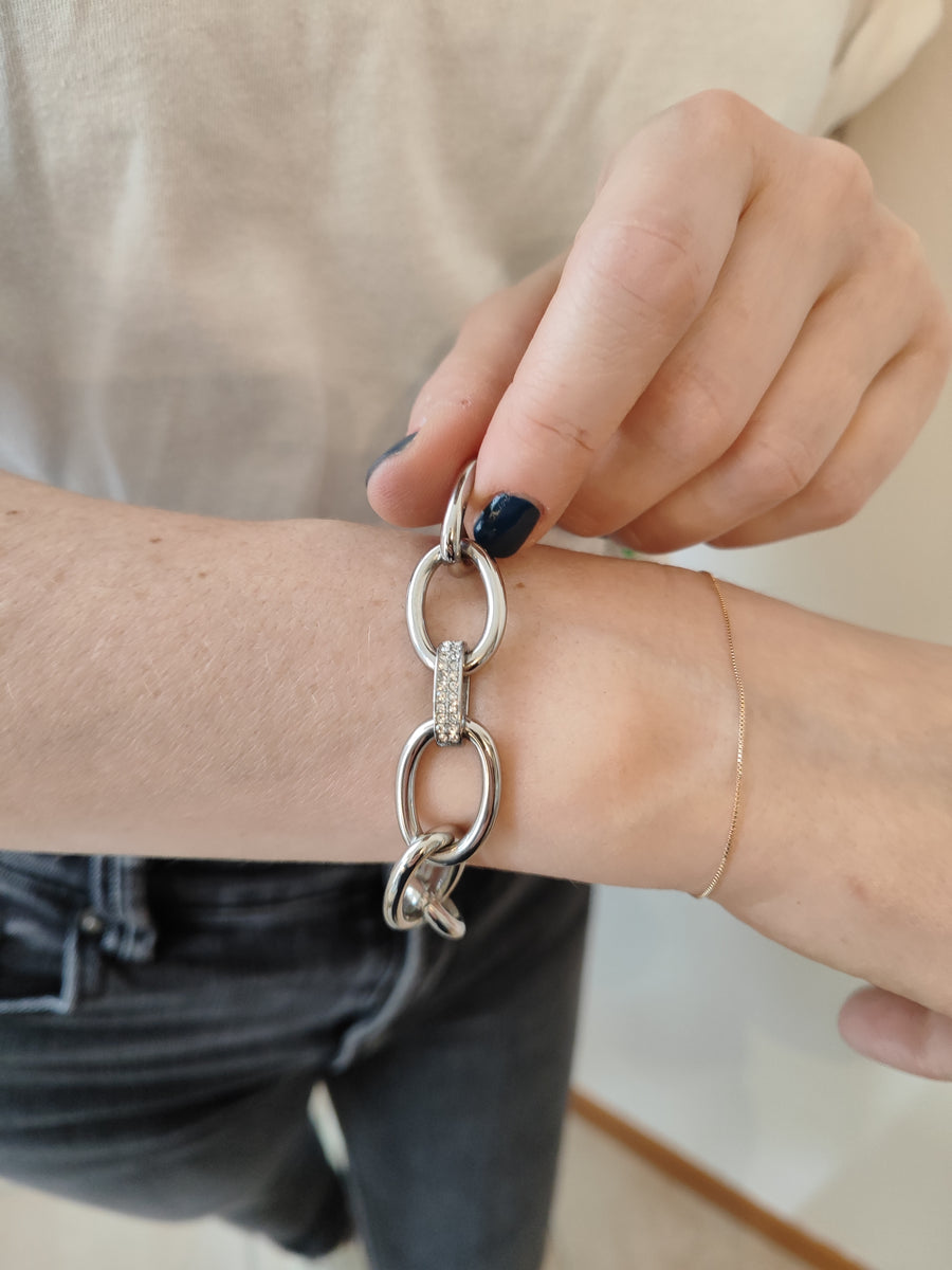 Bracciale Sandra in acciaio inossidabile argento, con maglie larghe e raccordo centrale decorato con strass. È indossato da una modella con maglia bianca e jeans neri mentre lo sorregge con la mano.