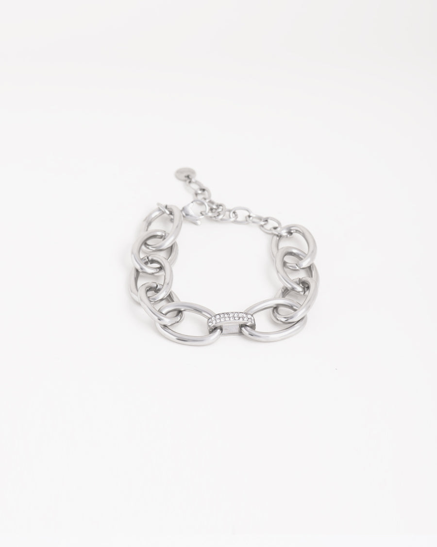 Bracciale Sandra in acciaio inossidabile a maglie larghe ovali color argento, con raccordo centrale a doppia fila di strass. È adagiato chiuso su uno sfondo chiaro. Come sempre è in materiale anallergico e waterproof. La chiusura è a moschettone con catenella per la regolazione.