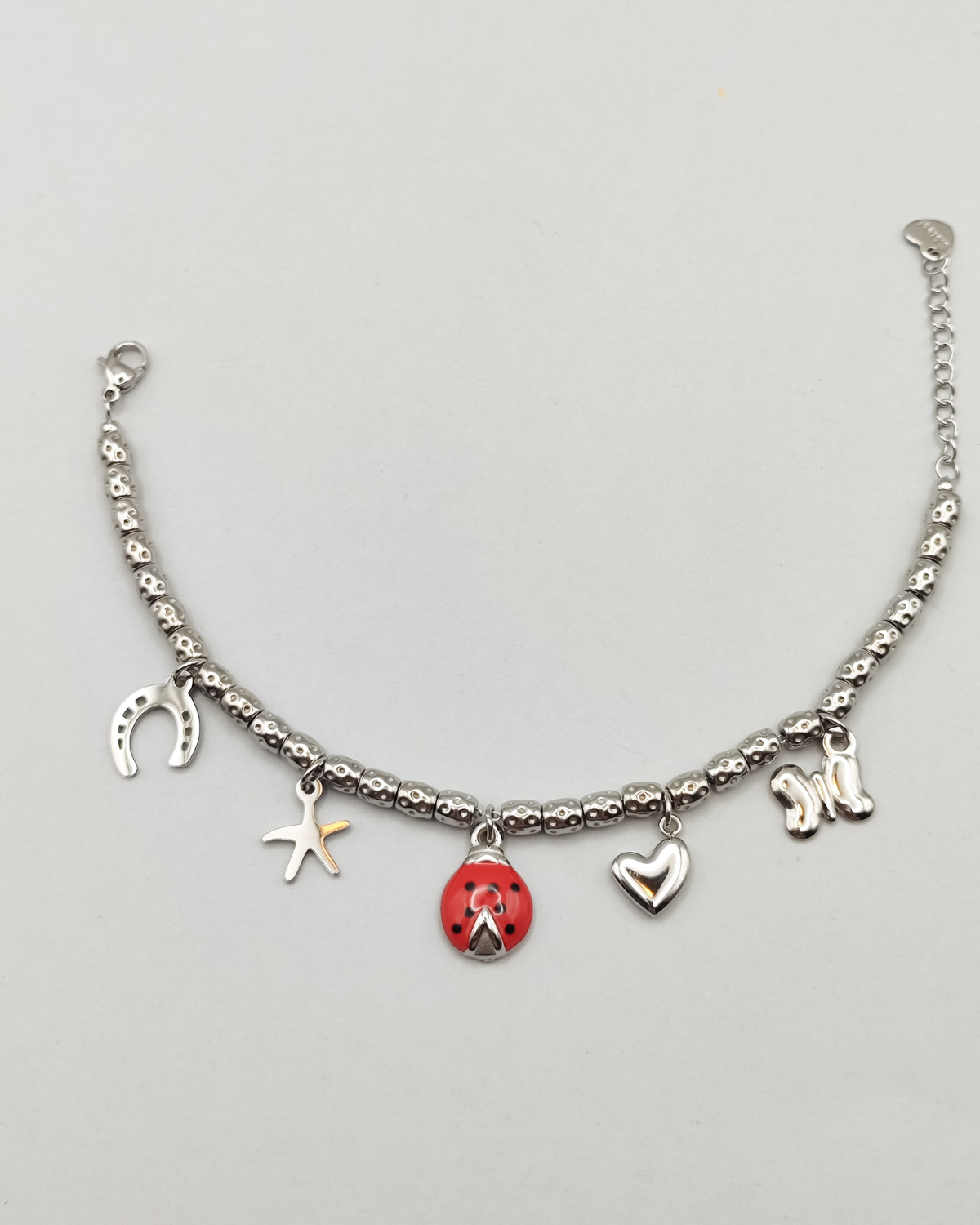 Bracciale Sara in acciaio inossidabile anallergico color argento con charms a forma di ferro di cavallo, stella, coccinella smaltata rossa, cuore e farfalla. Il bracciale è composto da perline martellate. È rappresentato aperto.