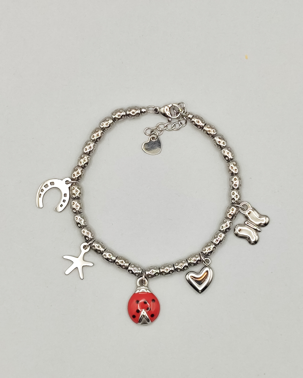 Bracciale Sara in acciaio inossidabile anallergico color argento con charms a forma di ferro di cavallo, stella, coccinella smaltata rossa, cuore e farfalla. Il bracciale è composto da perline martellate.