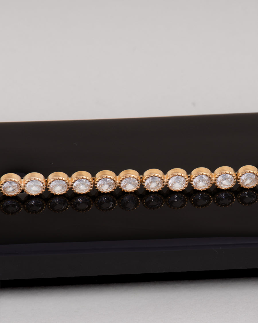 Bracciale Ylenia in acciaio inossidabile, anallergico e waterproof, modello tennis con strass montati su castoni rotondi zigrinati. È poggiato lateralmente su una superficie nera che lo riflette e il resto dello sfondo è bianco. 