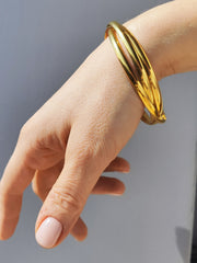 Bracciale Paola in acciaio inossidabile color oro. È ripreso indossato e si nota l'intreccio dei tre bracciali. La mano che lo porta è di fronte ad uno sfondo grigio.