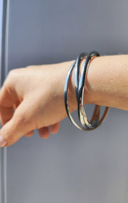 Bracciale Paola in versione argento indossato da una mano femminile su uno sfondo grigio. Si nota il design composto da tre singoli bracciali intrecciati tra loro.