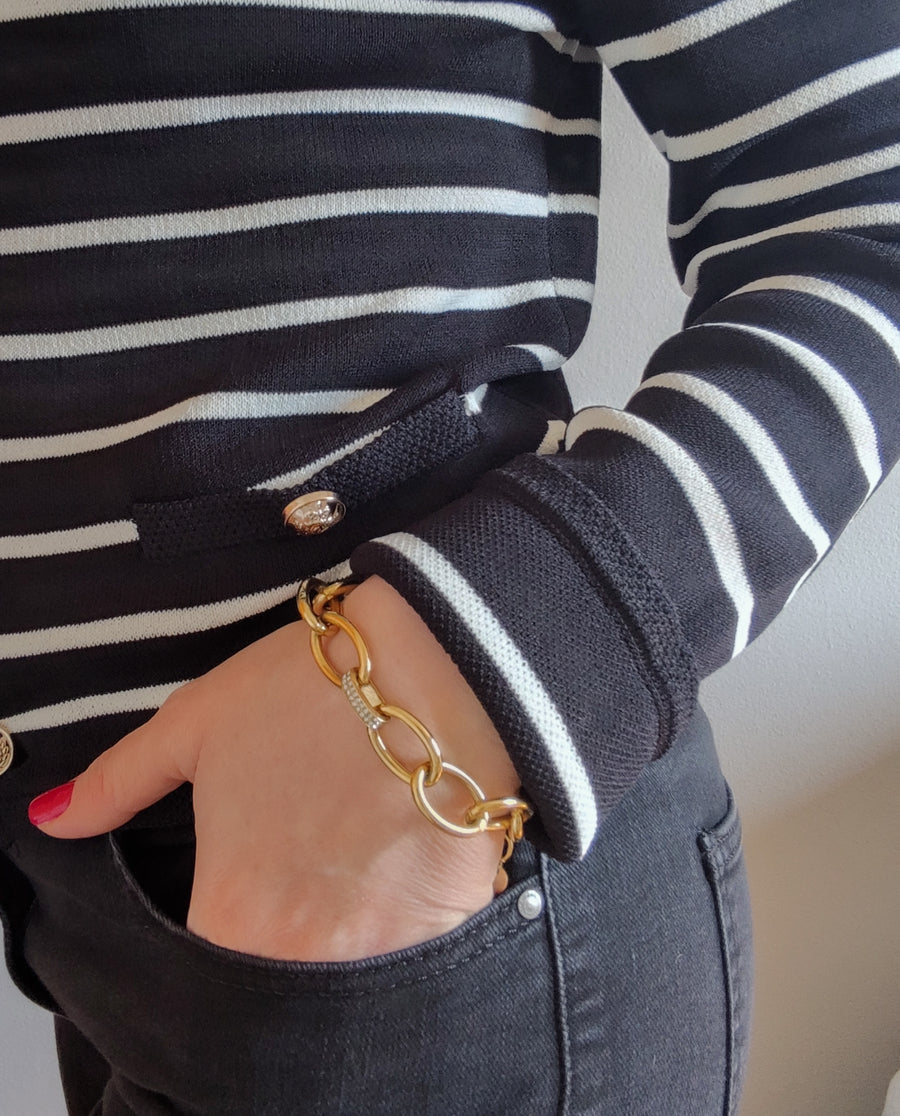 Bracciale Sandra in acciaio inossidabile color oro, indossato da una modella con cardigan a righe bianche e nere che tiene la mano in tasca. Il design del bracciale è a catena con maglie grandi e una maglia centrale con strass.