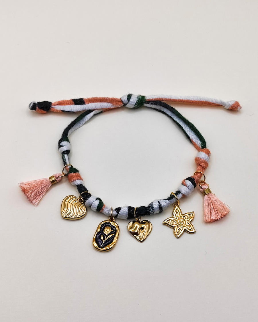 Cavigliera Isabel in fettuccia multicolor con nappine rosa e charms in acciaio. I charms rappresentano un cuore striato, una medaglietta smaltata nera con fiore in rilievo. un cuore sbalzato e una stella marina.