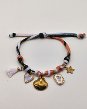 Cavigliera Isabel in fettuccia multicolor con nappina bianca e charms in acciaio inossidabile. I charms rappresentano un cuore smaltato bianco, una conchiglia, una medaglietta smaltata bianca con fiore in rilievo e una stella marina