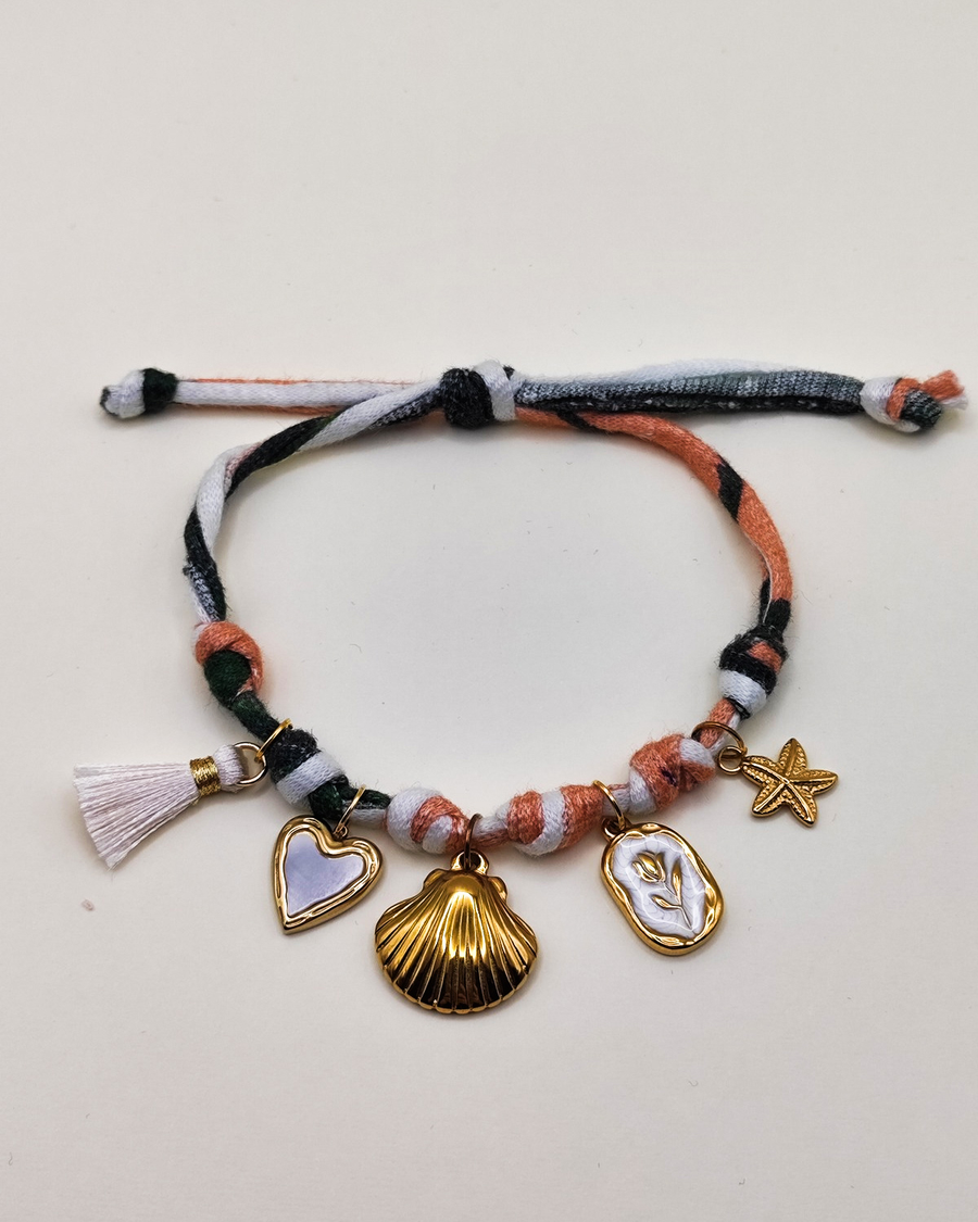 Cavigliera Isabel in fettuccia multicolor con nappina bianca e charms in acciaio inossidabile. I charms rappresentano un cuore smaltato bianco, una conchiglia, una medaglietta smaltata bianca con fiore in rilievo e una stella marina