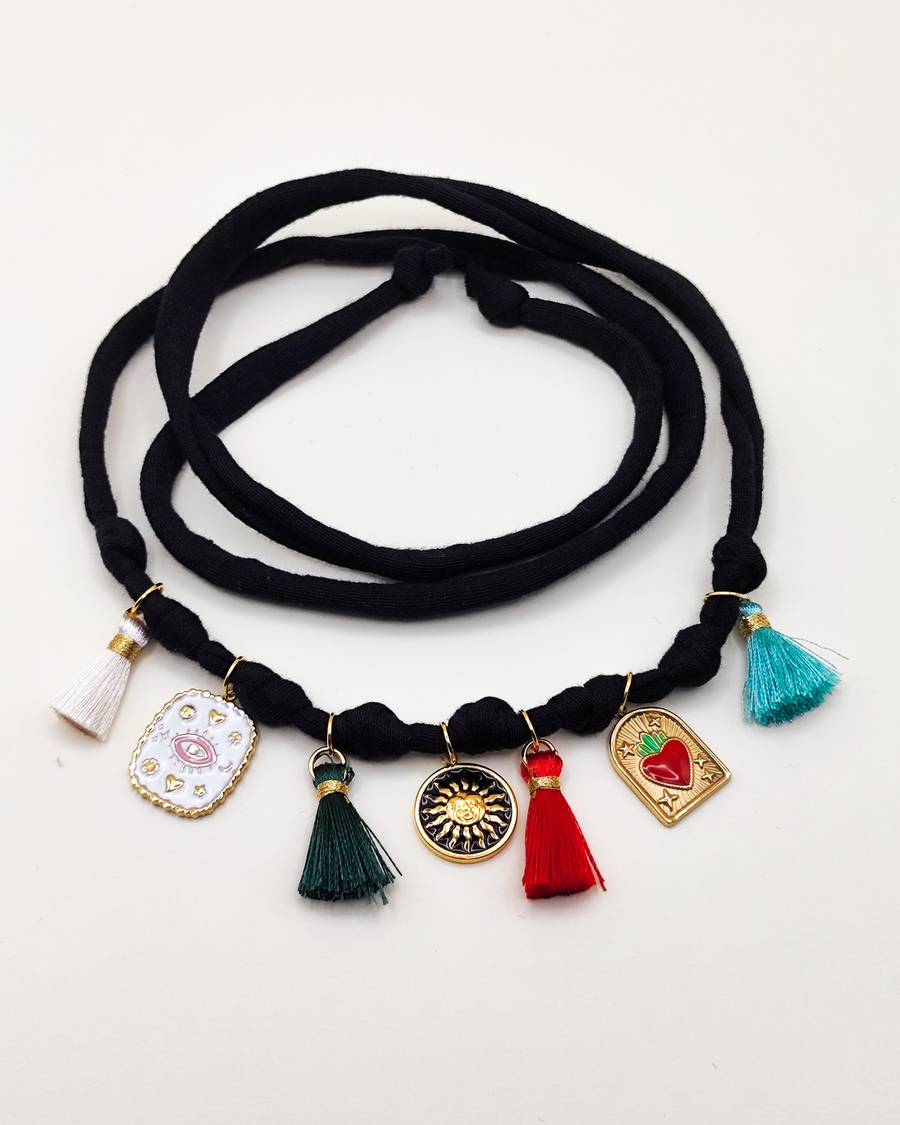Cavigliera Isabel in fettuccia elasticizzata con nappine multicolor e charms in acciaio inossidabile. I charms  rappresentano una medaglietta smaltata bianca con occhio rosa, una medaglietta tonda smaltata nera con un sole e una medaglietta on cuore sacro rosso e verde 