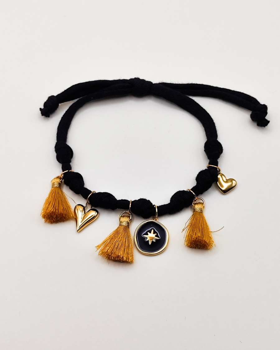 Cavigliera Isabel in fettuccia nera elasticizzata con nappine dorate e charms in acciaio. I charms rappresentano due cuori di diverse dimensioni alle estremità e centralmente una medaglietta smaltata nera con una stella dorata in rilievo