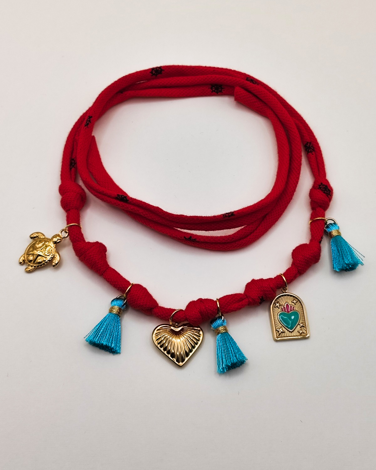 Caviglierea Isabel in fettuccia di cotone con nappine azzurre e charms in acciaio inossidabile. I charms rappresentano una tartaruga, un cuore striato e una medaglietta con cuore sacro smaltato azzurro e fuxia.