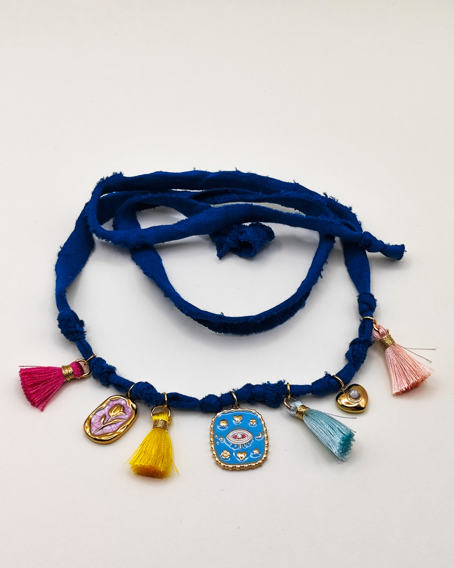 Cavigliera Isabel in fettuccia color ottanio con nappine multicolor e charms in acciaio inossidabile. I charms sono rappresentati da una medaglietta smaltata rosa con un fiore in rilievo, una medaglietta smaltata azzurra con un occhio e un pendende a cuore con brillantino al centro.