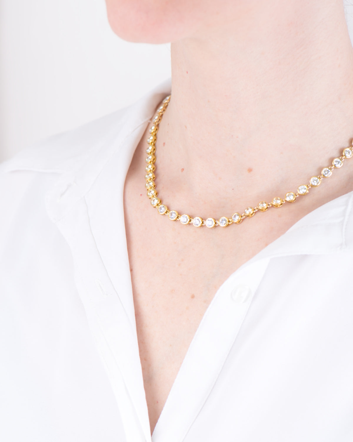 Collana Alessia in acciaio inossidabile color oro, modello tennis con strass indossata da una modella con camicia bianca. I castoni hanno una forma rotonda e sono uniti da una singola maglia. E'un modello a girocollo che però si può allungare.