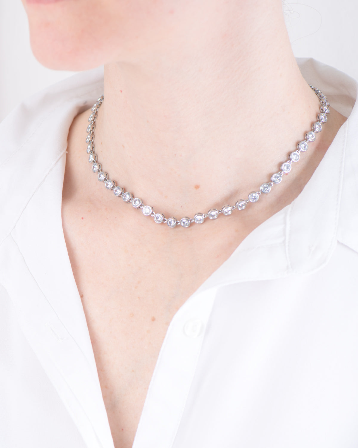 Collana Alessia in acciaio inossidabile color argento, modello tennis con strass indossata da una modella con camicia bianca. I castoni hanno una forma rotonda e sono uniti da una singola maglia. E'un modello a girocollo che però si può allungare.