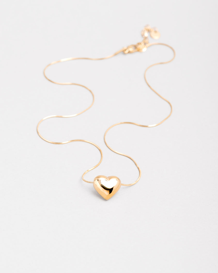 Collana Jessica in acciaio inossidabile color oro. È stesa su uno sfondo chiaro, la catena sottile è posizionata in modo sinuoso ed al centro c'è un cuore lucido bombato.