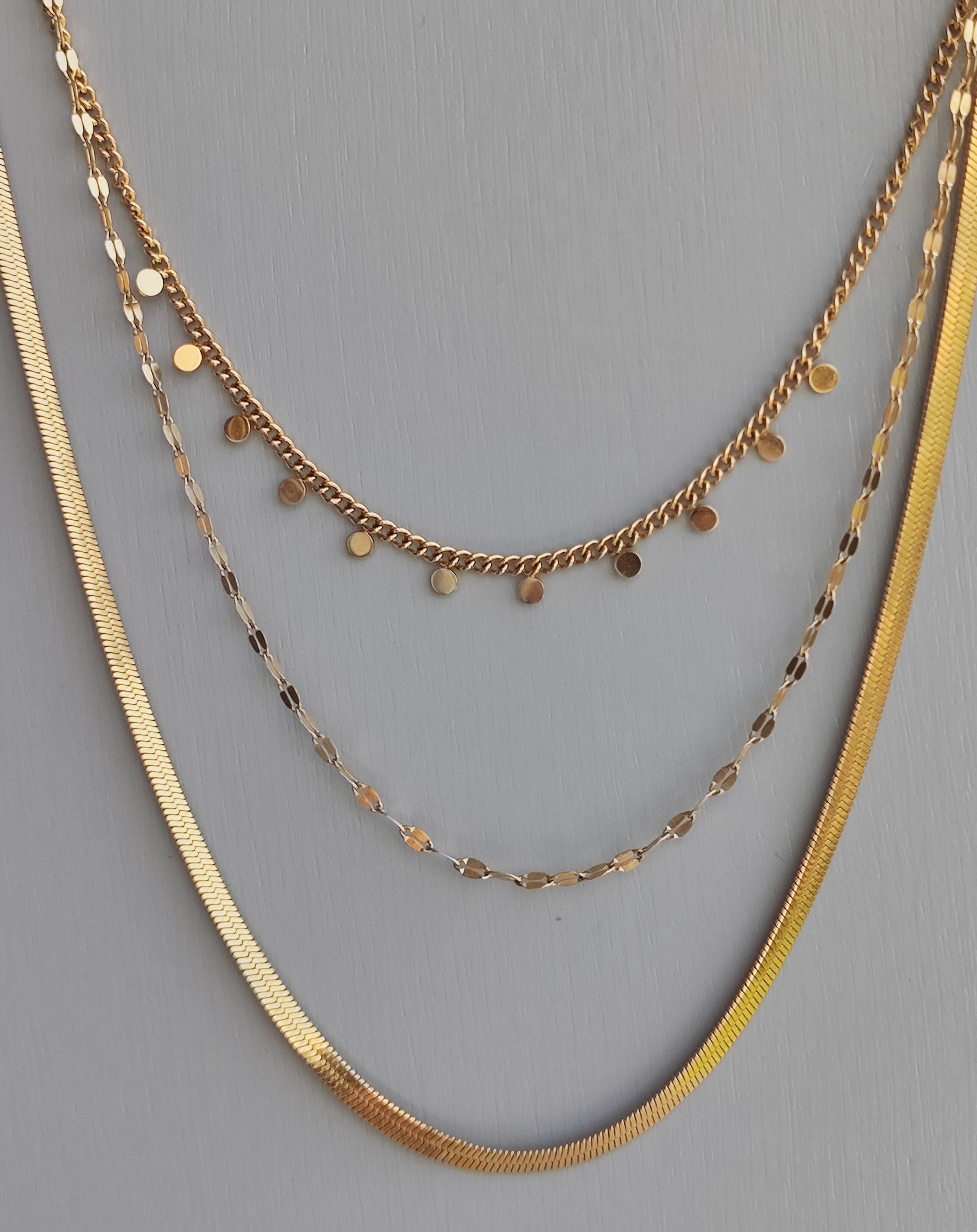 Collana Moira in acciaio inossidabile color oro. È ripresa frontalmente su uno sfondo grigio. È composta da tre fili di diversa lunghezza: il primo, corto, ha delle piccole medagliette rotonde, il secondo è una catena dalle maglie piatte e allungate, la terza è un modello snake