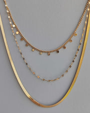 Collana Moira in acciaio inossidabile color oro. È ripresa frontalmente su uno sfondo grigio. È composta da tre fili di diversa lunghezza: il primo, corto, ha delle piccole medagliette rotonde, il secondo è una catena dalle maglie piatte e allungate, la terza è un modello snake