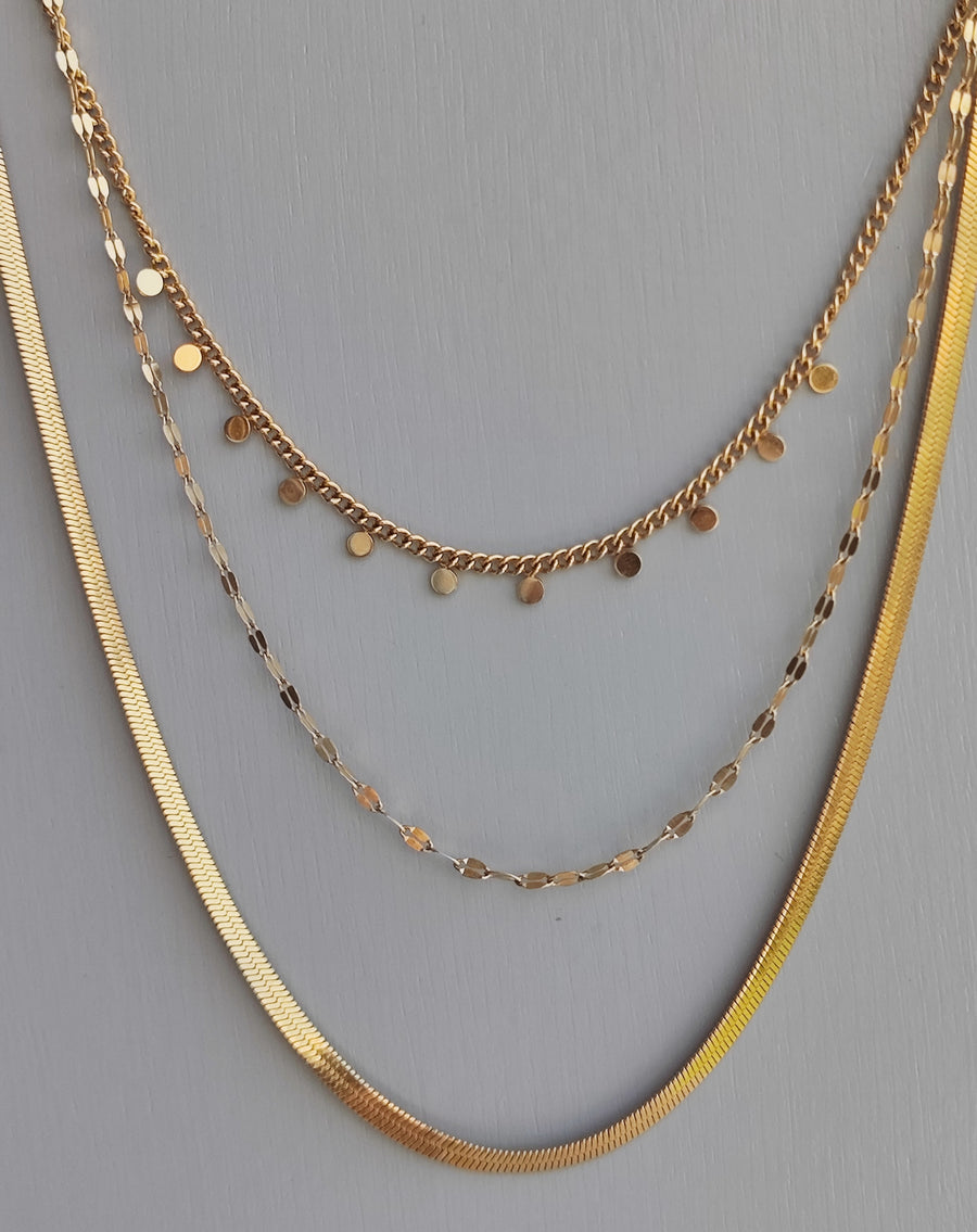 Collana Moira in acciaio inossidabile color oro. È ripresa frontalmente su uno sfondo grigio. È composta da tre fili di diversa lunghezza: il primo, corto, ha delle piccole medagliette rotonde, il secondo è una catena dalle maglie piatte e allungate, la terza è un modello snake