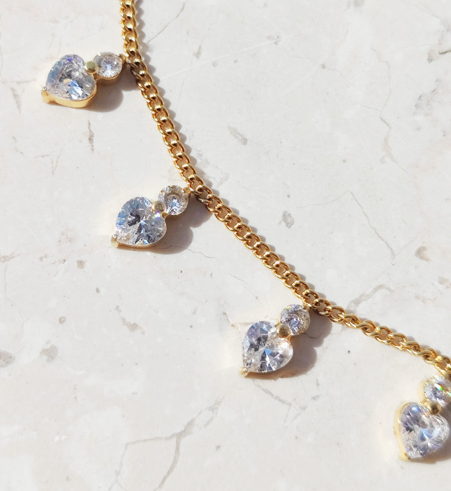 Dettaglio della collana Stefania in acciaio inossidabile color oro. Si vedono i cuoricini in strass sormontati da un brillantino, poggiati su una superficie di marmo e la maglia della catenina.