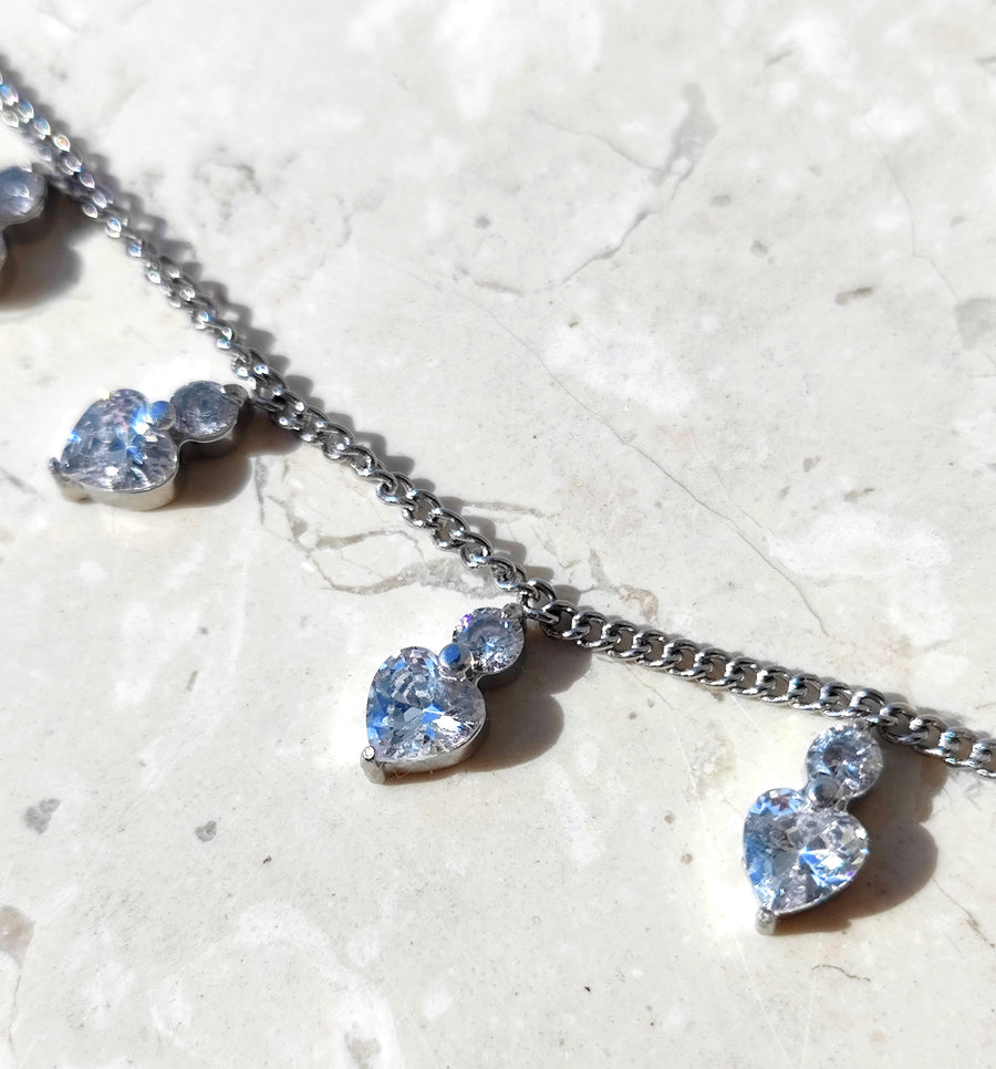 Dettaglio della collana Stefania in acciaio inossidabile color silver. Si vedono i cuoricini in strass sormontati da un brillantino, poggiati su una superficie di marmo e la maglia della catenina.