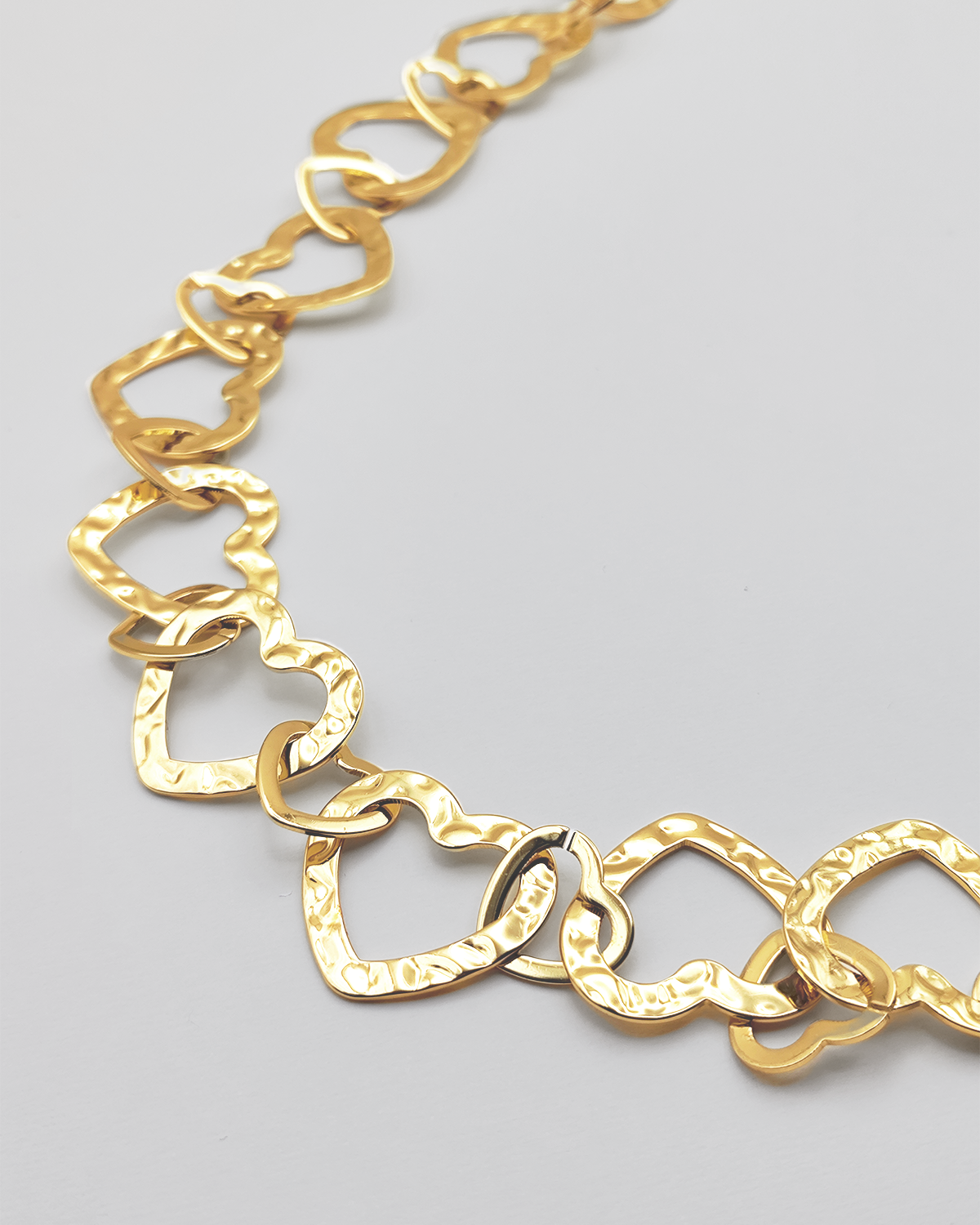 Dettaglio della collana Teresa in color oro. E' formata da tanti cuori in acciaio martellato intrecciati tra loro. E' ripresa su uno sfondo grigio chiaro.