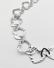 Dettaglio della collana Teresa in color argento. E' formata da tanti cuori in acciaio martellato intrecciati tra loro. E' ripresa su uno sfondo grigio chiaro.