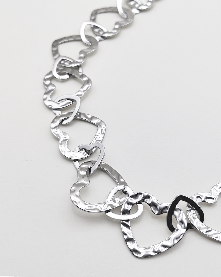 Dettaglio della collana Teresa in color argento. E' formata da tanti cuori in acciaio martellato intrecciati tra loro. E' ripresa su uno sfondo grigio chiaro.