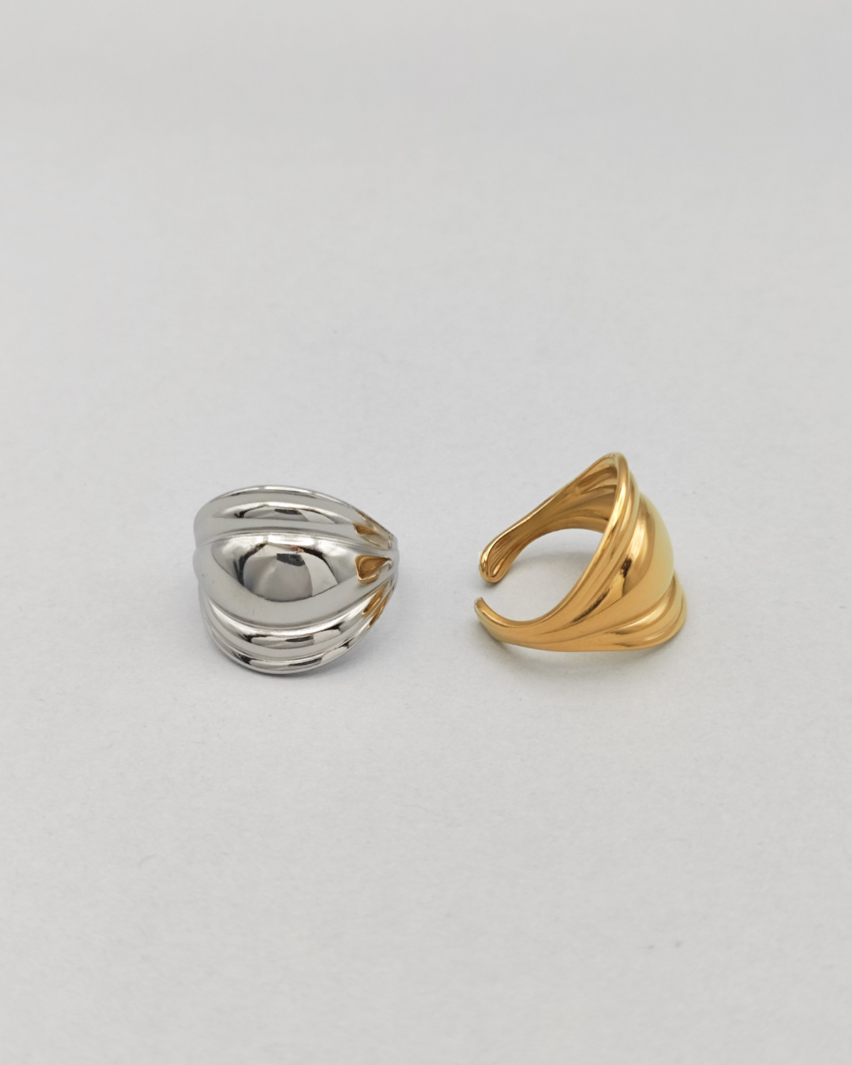 Coppia di anelli Carlotta in acciaio inossidabile anallergico color argento e oro. L'anello argento èrappresentato frontalmente su uno sfondo grigio chiaro. Ha una forma bombata al centro che si ristringe verso i lati ed ha un finish lucido e si vede la chiusura regolabile sul retro. Quello dorato è invece ripreso lateralmente.