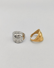 Coppia di anelli Carlotta in acciaio inossidabile anallergico color argento e oro. L'anello argento èrappresentato frontalmente su uno sfondo grigio chiaro. Ha una forma bombata al centro che si ristringe verso i lati ed ha un finish lucido e si vede la chiusura regolabile sul retro. Quello dorato è invece ripreso lateralmente.