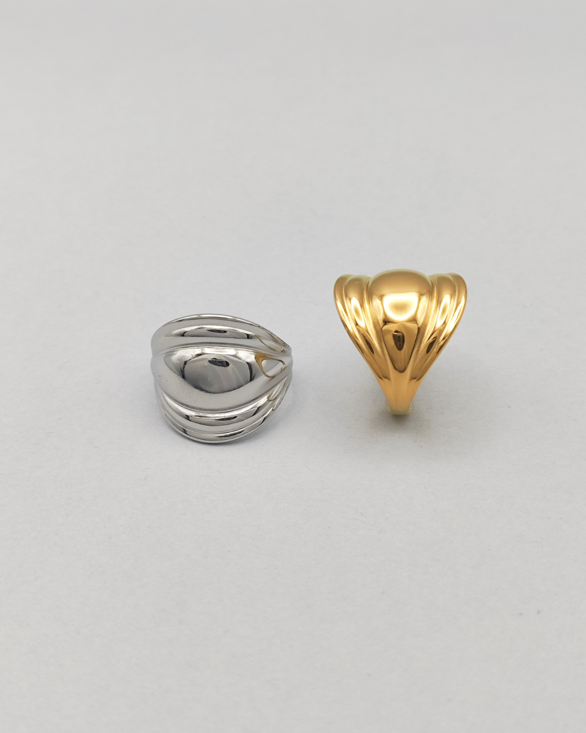 Coppia di anelli Carlotta in acciaio inossidabile anallergico color argento e oro. L'anello argento èrappresentato frontalmente su uno sfondo grigio chiaro. Ha una forma bombata al centro che si ristringe verso i lati ed ha un finish lucido e si vede la chiusura regolabile sul retro. Quello dorato è invece ripreso lateralmente.