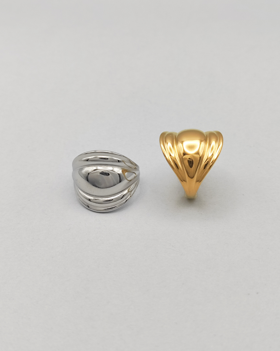 Coppia di anelli Carlotta in acciaio inossidabile anallergico color argento e oro. L'anello argento èrappresentato frontalmente su uno sfondo grigio chiaro. Ha una forma bombata al centro che si ristringe verso i lati ed ha un finish lucido e si vede la chiusura regolabile sul retro. Quello dorato è invece ripreso lateralmente.